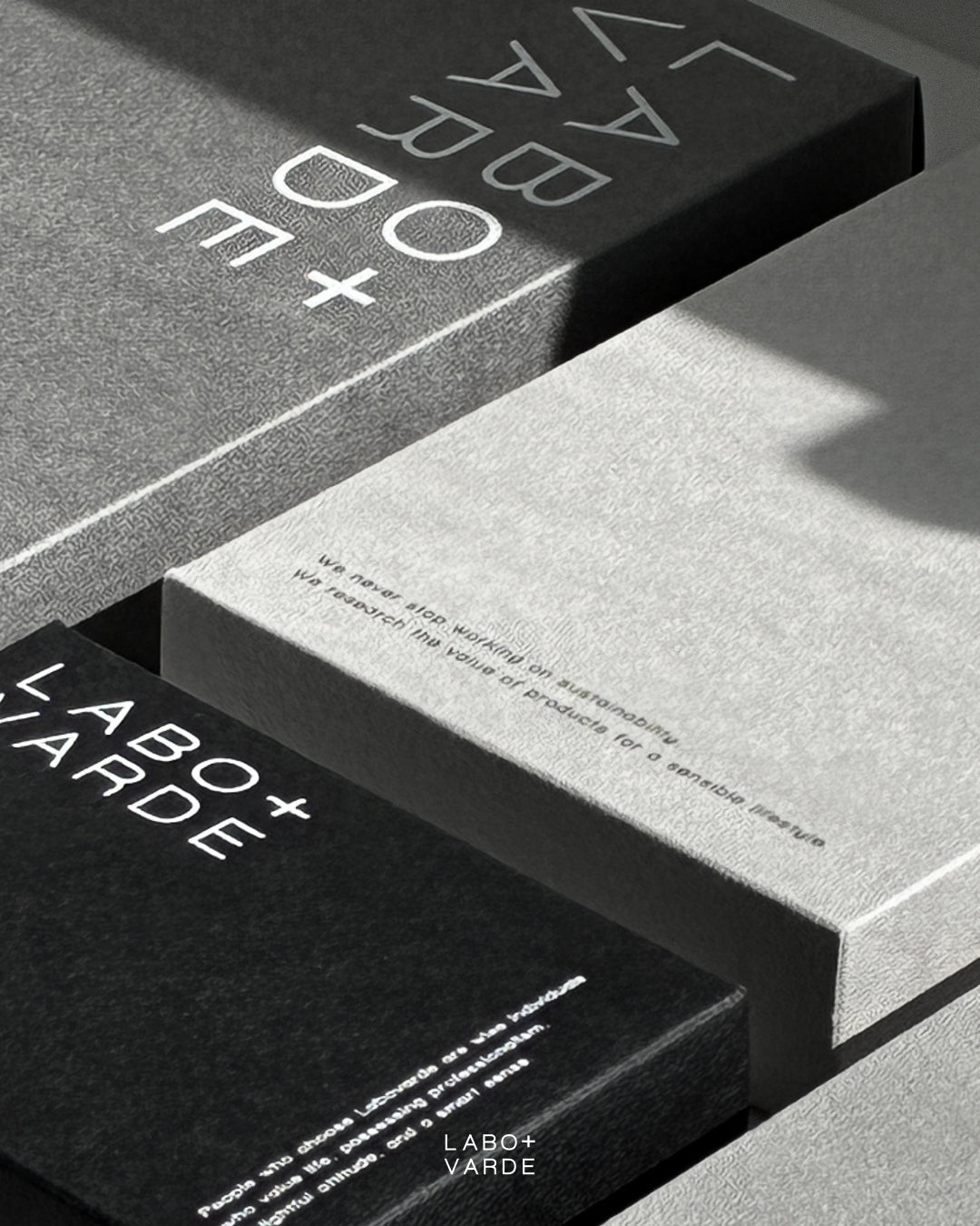 Close-up of minimalist gray and black Labovarde eco-friendly paper packaging boxes, featuring printed brand typography about sustainability and the LABO+VARDE logo under elegant diagonal shadows.